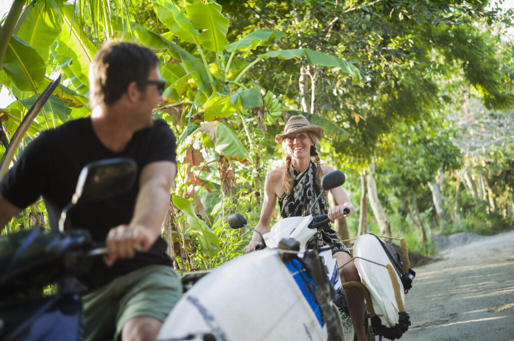 Een stel rijdt op een scooter in Ubud, Bali, terwijl ze een surfplank dragen.