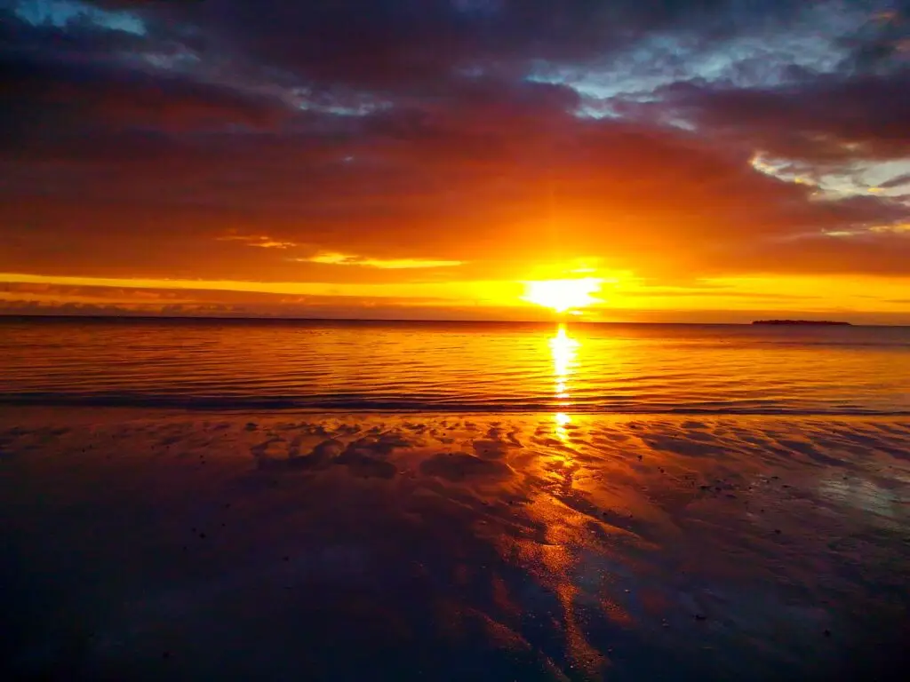 The sun sets over the beach with bright orange and yellow colors reflected in the wet sand.